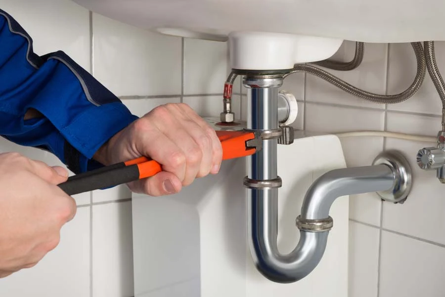 Plombier en combinaison bleue utilisant une cl&eacute; &agrave; molette avec manches orange et noir pour serrer un raccord de siphon sous un &eacute;vier