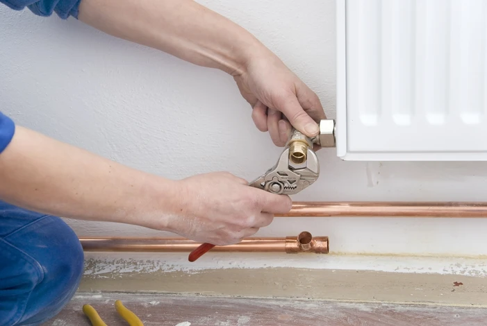 Plombier en combinaison bleue travaillant à genoux sur raccordement de tuyauterie cuivre au pied d'un radiateur blanc