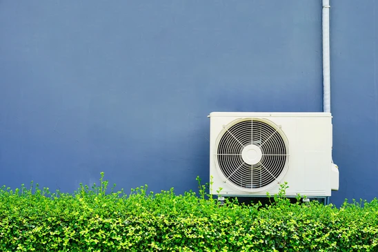 Sc&egrave;ne ext&eacute;rieure avec unit&eacute; de climatisation blanche install&eacute;e sur support m&eacute;tallique contre mur bleu, entour&eacute;e de v&eacute;g&eacute;tation verte dense