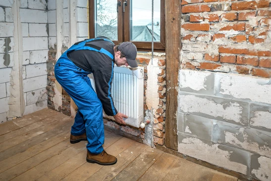Ouvrier en salopette bleue installant un radiateur blanc contre un mur en briques apparentes dans une maison en r&eacute;novation