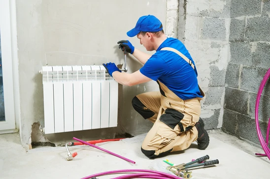 Ouvrier en salopette bleue installant un radiateur blanc contre un mur en briques apparentes dans une maison en rénovation