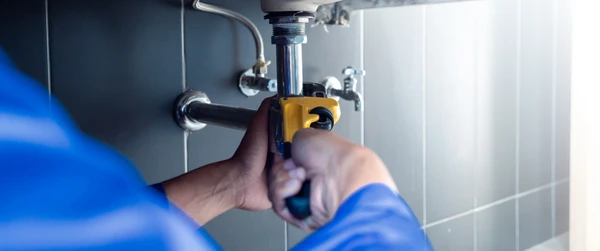Plombier en uniforme professionnel bleu intervenant à domicile, diagnostiquant une fuite d'eau au niveau d'une tuyauterie en cuivre visible sur un mur. Le plombier tient un équipement de diagnostic ou de détection de fuite, dans un intérieur résidentiel, illustrant clairement l'action de dépannage d'urgence et de diagnostic. L'image doit montrer la réactivité et la compétence du professionnel face à une situation d'urgence plomberie.