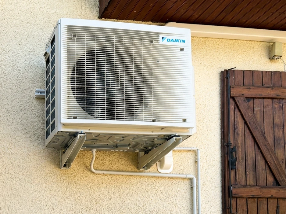 Unité extérieure de climatisation **Daikin** fixée en hauteur sur un mur de façade crépi, sous un auvent à charpente bois