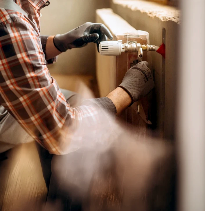 Homme en chemise à carreaux effectuant des travaux de plomberie sur un radiateur blanc dans un intérieur résidentiel lumineux près d'une fenêtre